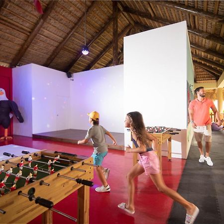 Impressive Punta Cana Esterno foto The photo shows an indoor recreational space with a thatched roof. In the foreground, there is a foosball table with figures attached to it, where two children are playing or running towards it. A man and a woman, likely parents, are walking together