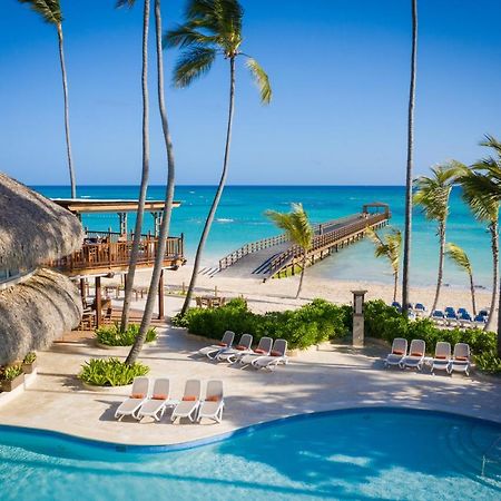 Impressive Punta Cana Esterno foto The photo depicts a serene tropical scene featuring a swimming pool in the foreground, surrounded by lounge chairs. In the background, there is a beautiful beach with clear turquoise waters and a wooden pier extending out into the ocean. Palm trees f