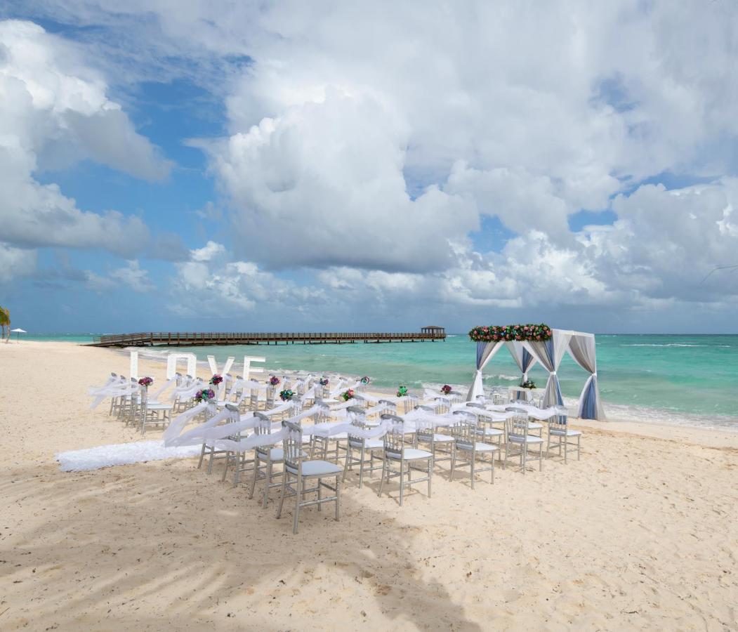 Impressive Punta Cana Esterno foto The photo shows a beach setting that appears to be prepared for a wedding ceremony. There are rows of white chairs arranged on the sand facing a decorated archway. The arch is draped in white fabric and adorned with floral elements. In the background