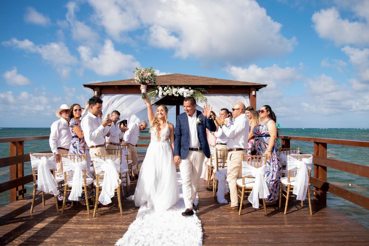 Impressive Punta Cana Esterno foto The photo shows a wedding ceremony taking place on a wooden pier over water. A bride and groom are at the center, with the bride holding her bouquet up in celebration. They are surrounded by a group of guests, all dressed in formal attire, some clapp