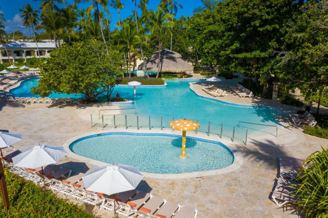 Impressive Punta Cana Esterno foto The photo shows a vibrant outdoor pool area surrounded by lush greenery. There are two distinct pool sections visible: a larger main pool with a clear blue water and a smaller, circular pool that features a playful mushroom-shaped fountain. Sun loung