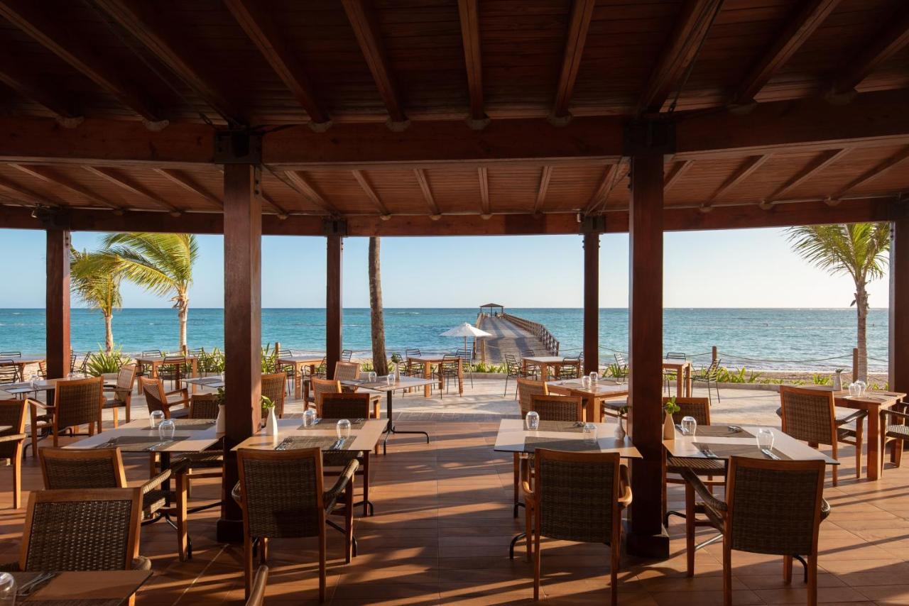 Impressive Punta Cana Esterno foto The photo shows a beachfront dining area with a wooden structure featuring a thatched roof. There are several tables and chairs arranged on a wooden deck, providing a picturesque view of the ocean. Palm trees can be seen swaying in the breeze, and th