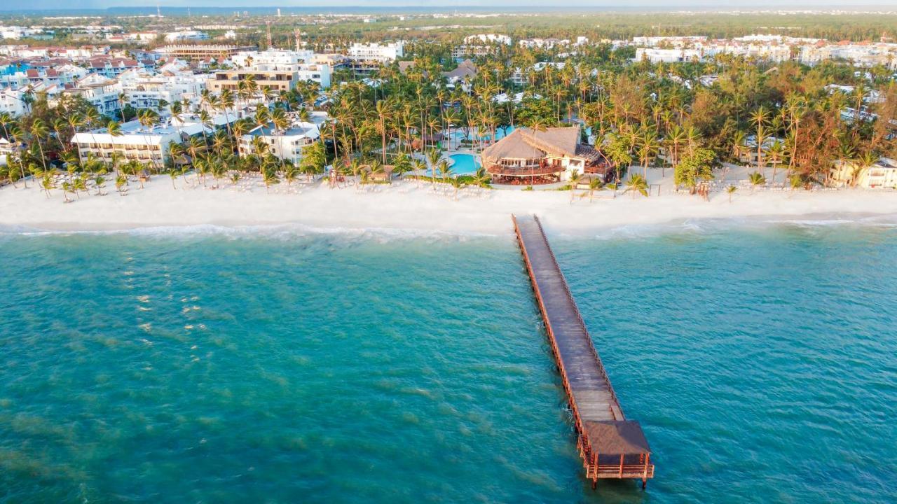 Impressive Punta Cana Esterno foto The photo shows a picturesque beach scene viewed from above. In the foreground, there is a wooden pier extending over clear turquoise water. The pier leads towards a coastal area with a sandy beach lined by lush palm trees. In the background, several
