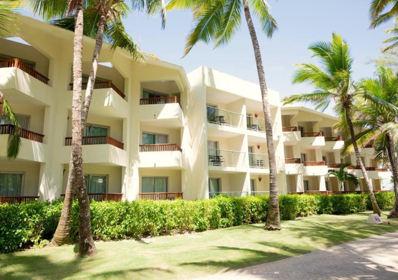Impressive Punta Cana Esterno foto The photo shows a beachfront hotel or resort building with multiple levels. The structure is light-colored with balconies that overlook a manicured garden area. Tall palm trees and lush green vegetation surround the building, suggesting a tropical lo