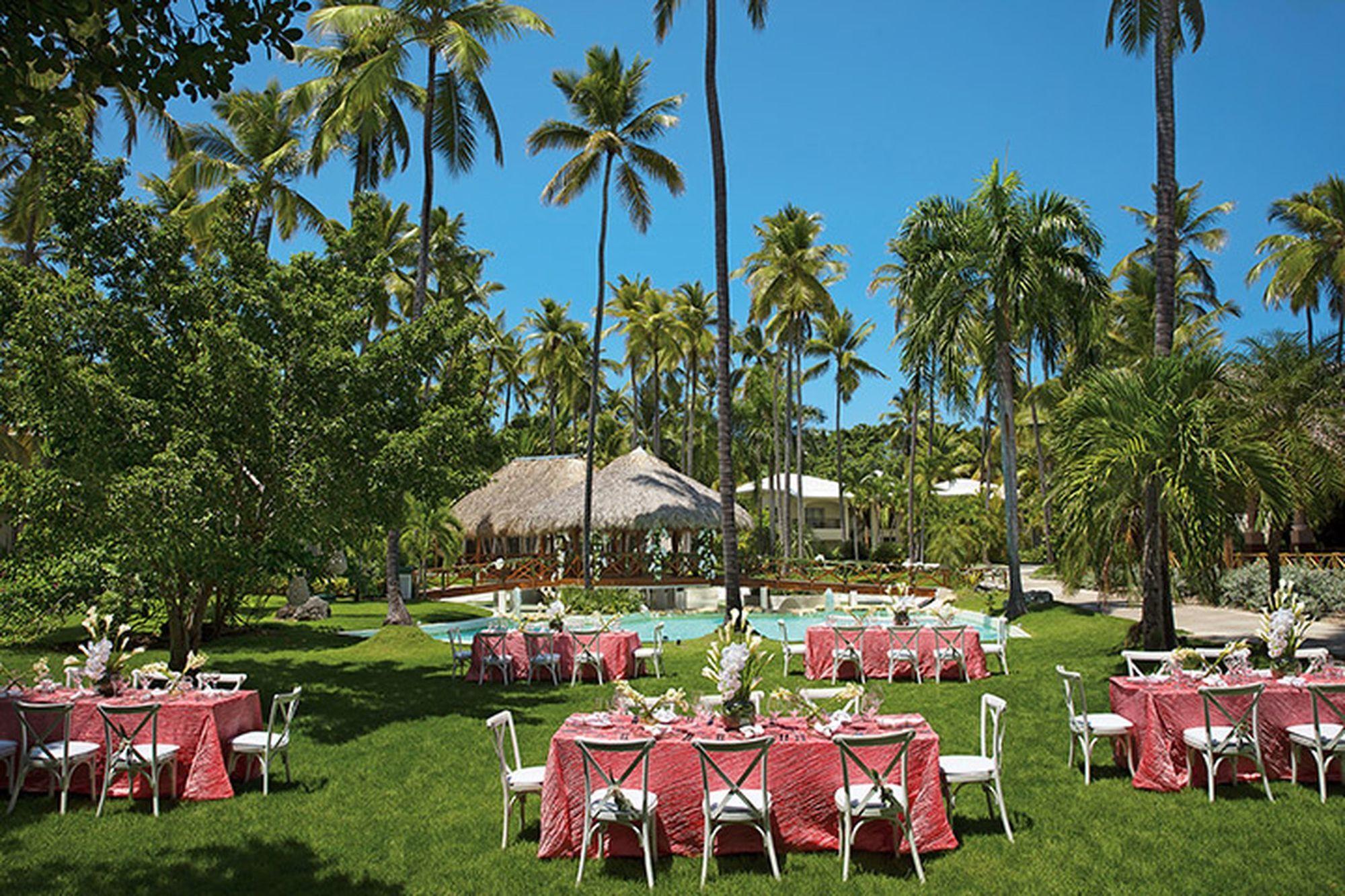 Impressive Punta Cana Esterno foto The photo depicts a beautiful outdoor setting, likely for an event or celebration. There are several tables arranged on a lush green lawn, each dressed with pink tablecloths. White chairs surround the tables, creating an inviting atmosphere. In the b