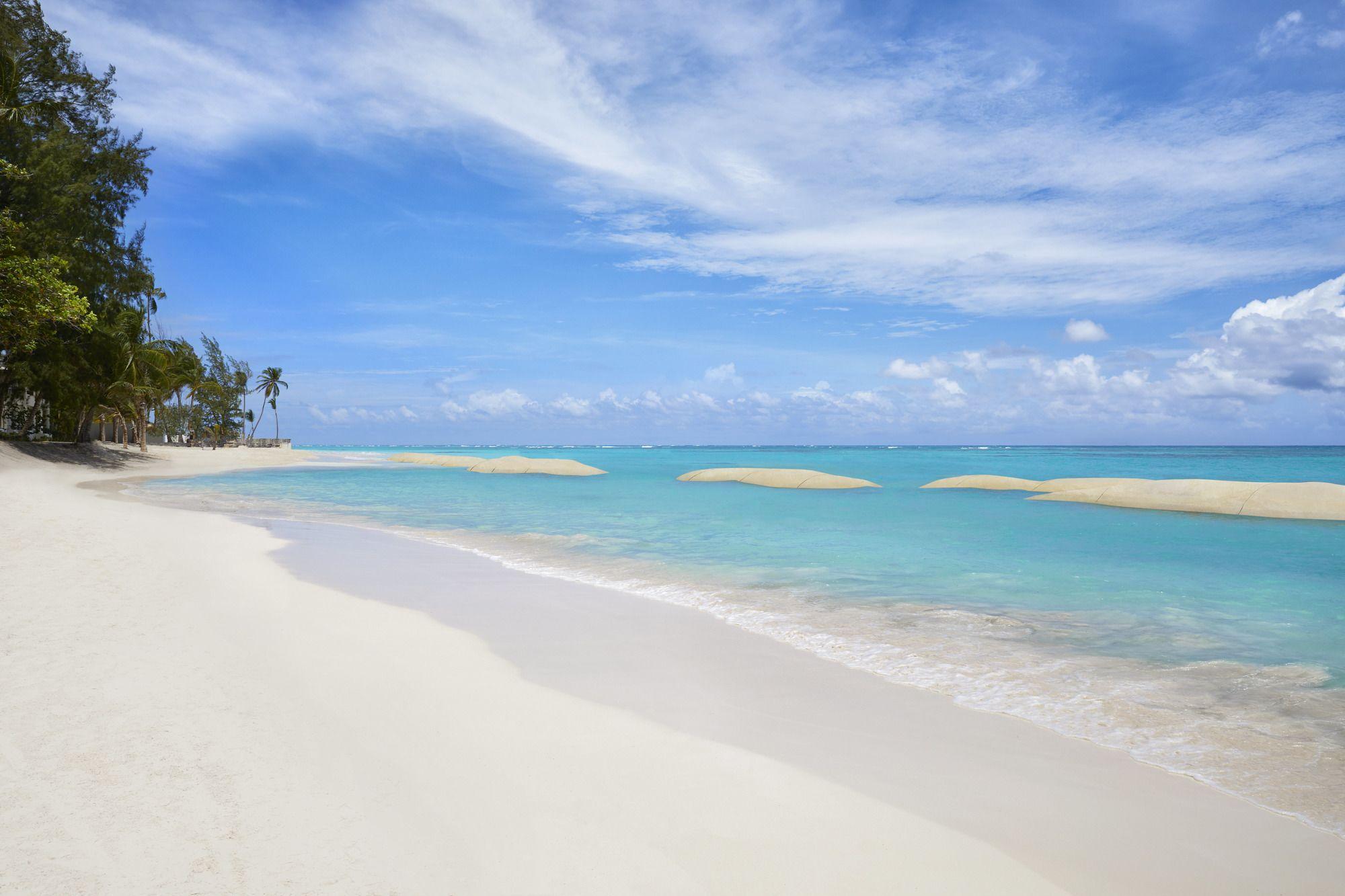 Impressive Punta Cana Esterno foto The photo showcases a beautiful beach scene. In the foreground, there is soft, white sand leading to clear, turquoise waters. Gentle waves lap at the shore. In the background, palm trees can be seen lining the beach, and the sky is bright with a few 