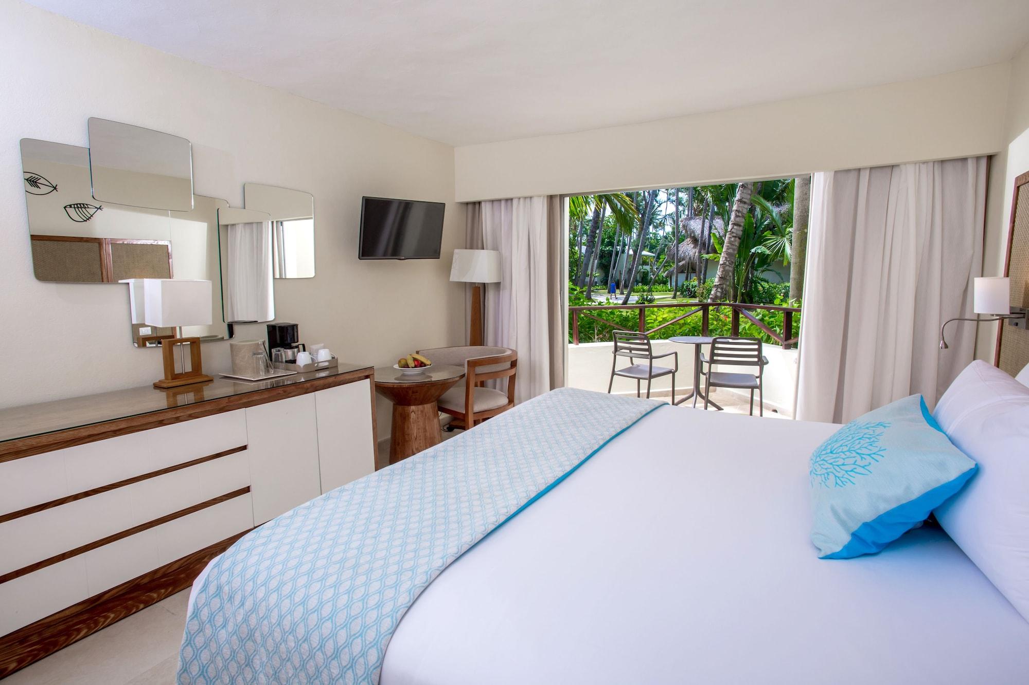 Impressive Punta Cana Esterno foto The photo shows a well-decorated hotel room featuring a large bed with a light blue throw blanket. There is a bedside table and a dresser with a mirror, as well as a television mounted on the wall. Natural light comes in through a large window that o