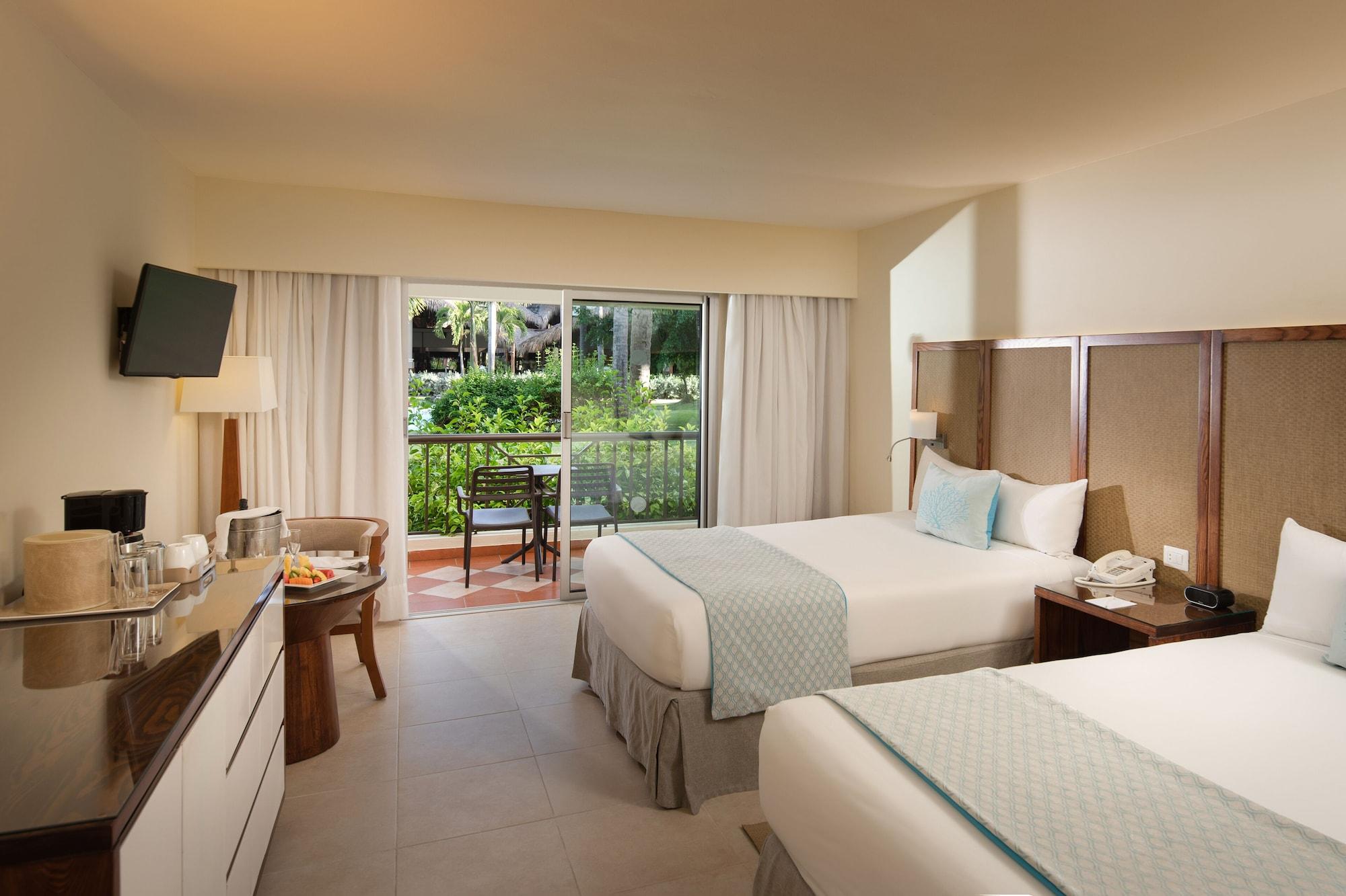 Impressive Punta Cana Esterno foto The photo shows a hotel room featuring two beds with light-colored linens and decorative pillows. There is a modern armchair beside a small table near a large window that offers a view of an outdoor garden area. A flat-screen TV is mounted on the wal