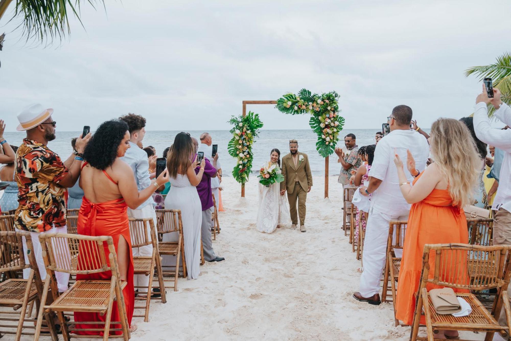 Impressive Punta Cana Esterno foto The photo depicts a wedding ceremony on a beach. In the center, there is an altar decorated with greenery and flowers. A couple is walking down the aisle, with guests on either side capturing the moment on their phones. The attendees are dressed in a