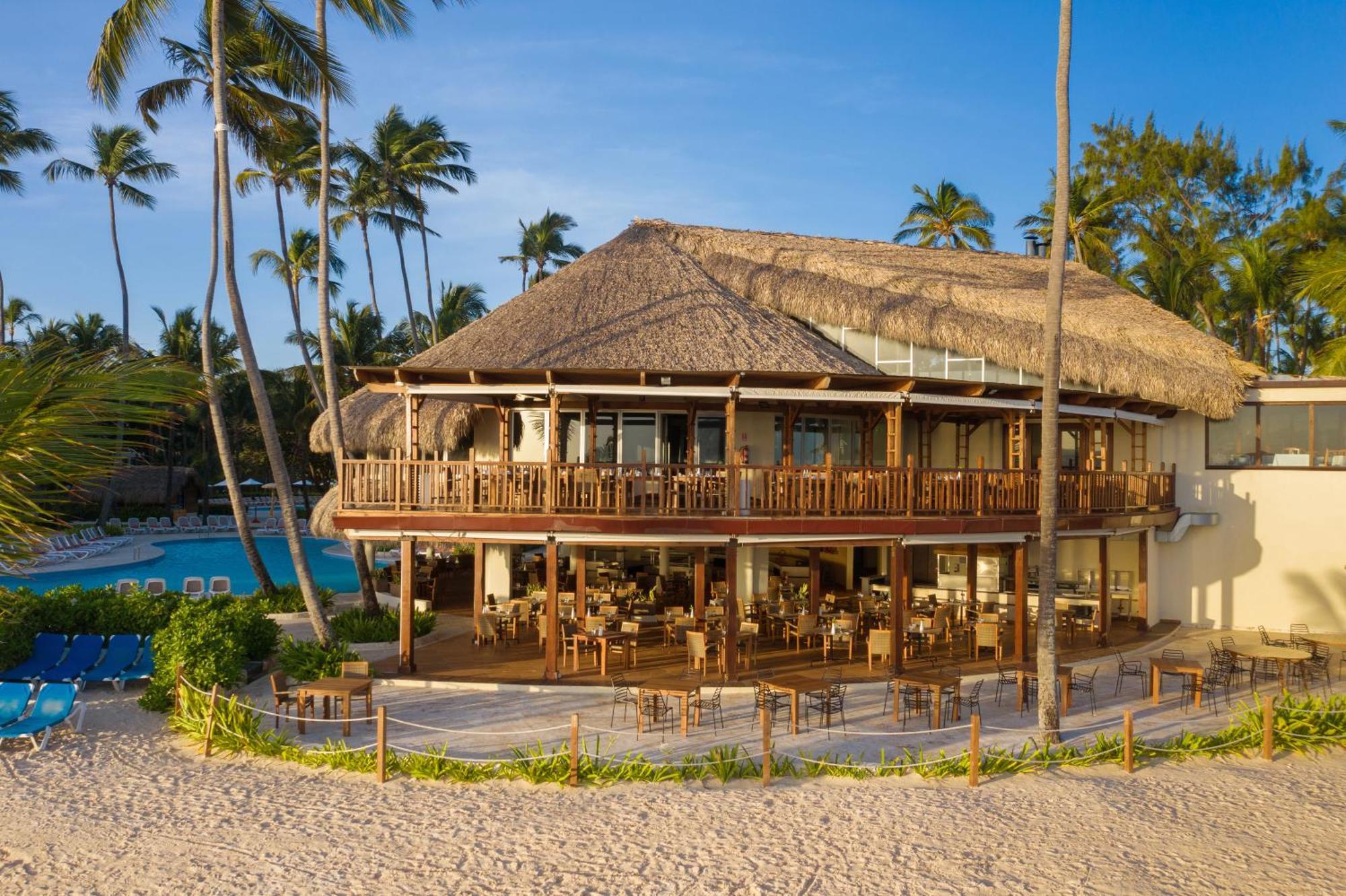 Impressive Punta Cana Esterno foto The photo shows a tropical beachfront building, likely a restaurant or resort, featuring a large deck with wooden railings and outdoor seating. The structure has a thatched roof and multiple large windows, allowing for a view of the surrounding area.