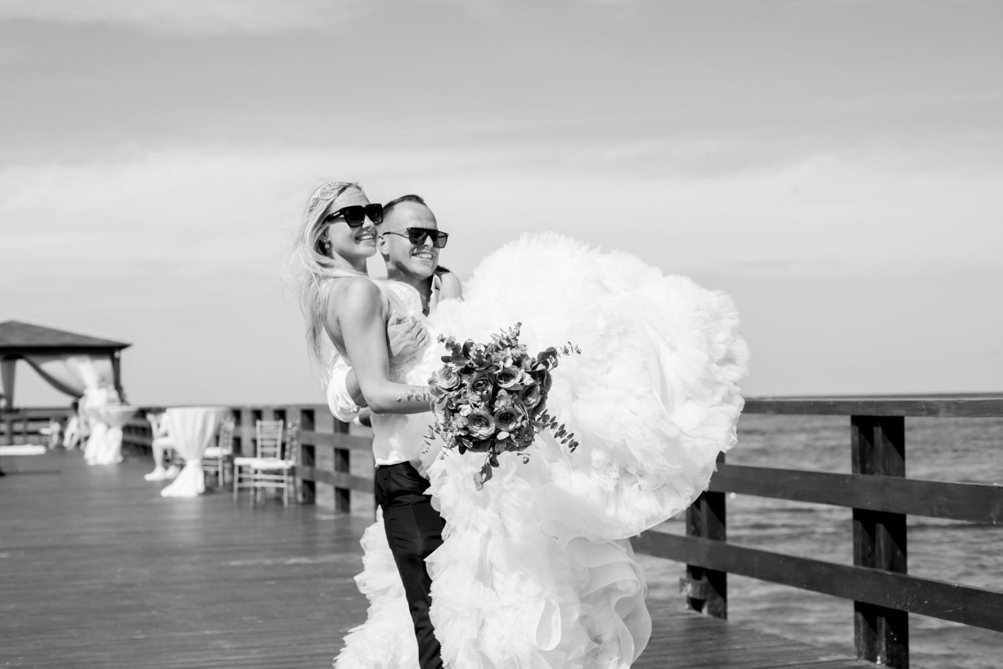 Impressive Punta Cana Esterno foto The photo appears to depict a couple celebrating their wedding. They are standing on a wooden pier by the water, with the ocean in the background. The scene is in black and white, which adds a classic touch. The bride is wearing a dramatic, fluffy we