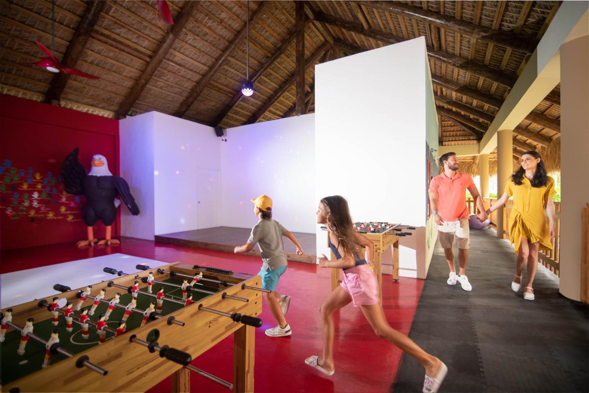 Impressive Punta Cana Esterno foto The photo shows an indoor recreational space with a thatched roof. In the foreground, there is a foosball table with figures attached to it, where two children are playing or running towards it. A man and a woman, likely parents, are walking together