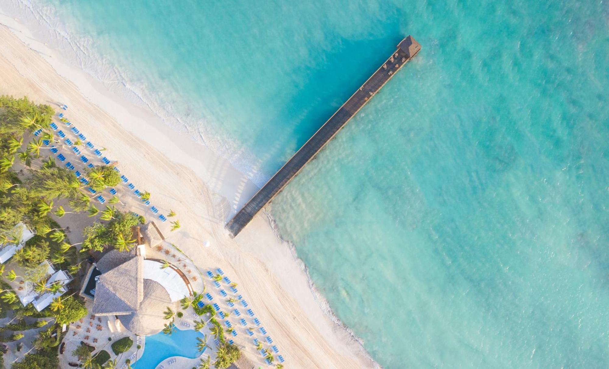 Impressive Punta Cana Esterno foto The photo shows an aerial view of a beautiful beach scene. There is a long wooden pier extending over clear turquoise waters, leading out from the sandy shore. The beach is lined with umbrellas or lounge chairs, and there is a large building with a t