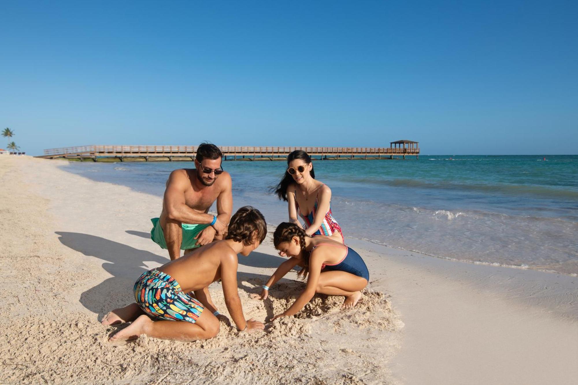 Impressive Punta Cana Esterno foto The photo depicts a family enjoying a sunny day at the beach. In the foreground, two children are playing in the sand, focused on digging or building something. One child is kneeling, while the other is sitting nearby. An adult male is watching them 