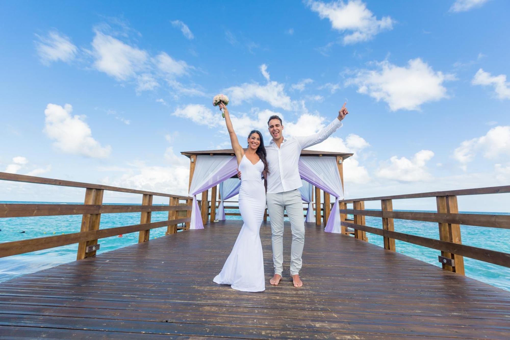 Impressive Punta Cana Esterno foto The photo shows a joyful couple standing on a wooden pier or deck over the water. The woman is wearing a beautiful white wedding dress and is holding a bouquet of flowers, while the man is dressed in light-colored attire, likely suitable for a beach 