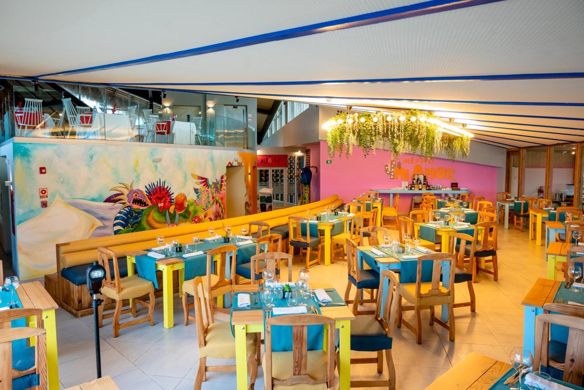 Impressive Punta Cana Esterno foto The photo shows a vibrant restaurant interior. It features wooden tables and chairs, painted in bright, cheerful colors like yellow and turquoise. The tables are set with utensils and blue tablecloths. The walls are decorated with colorful murals and