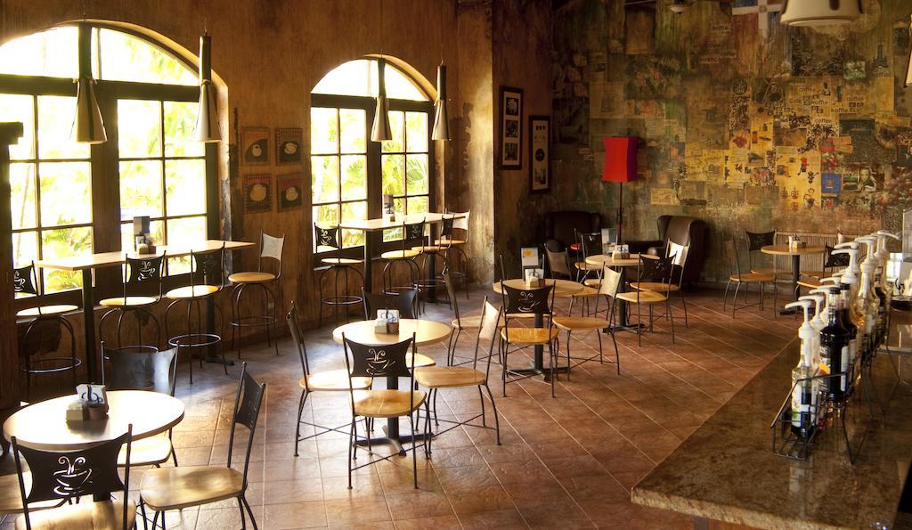Impressive Punta Cana Esterno foto The photo shows the interior of a café or restaurant. There are several small tables and chairs arranged throughout the space. Large windows allow natural light to fill the area, creating a warm atmosphere. The walls have a rustic, textured finish wi