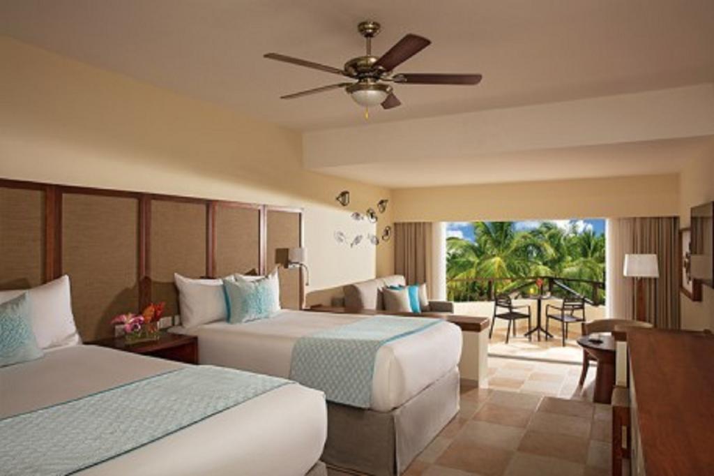 Impressive Punta Cana Esterno foto The photo shows a hotel room with two beds, decorated in a light, modern style. There is a ceiling fan above and large windows that open to a balcony. The room features soft colors, with a turquoise accent on the bedding, and has a small seating area