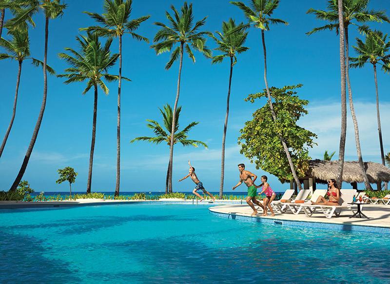 Impressive Punta Cana Esterno foto The photo showcases a vibrant tropical scene featuring a swimming pool surrounded by tall palm trees. In the foreground, several people are enjoying themselves, with one person jumping into the pool. Others are relaxing on lounge chairs by the poolsi