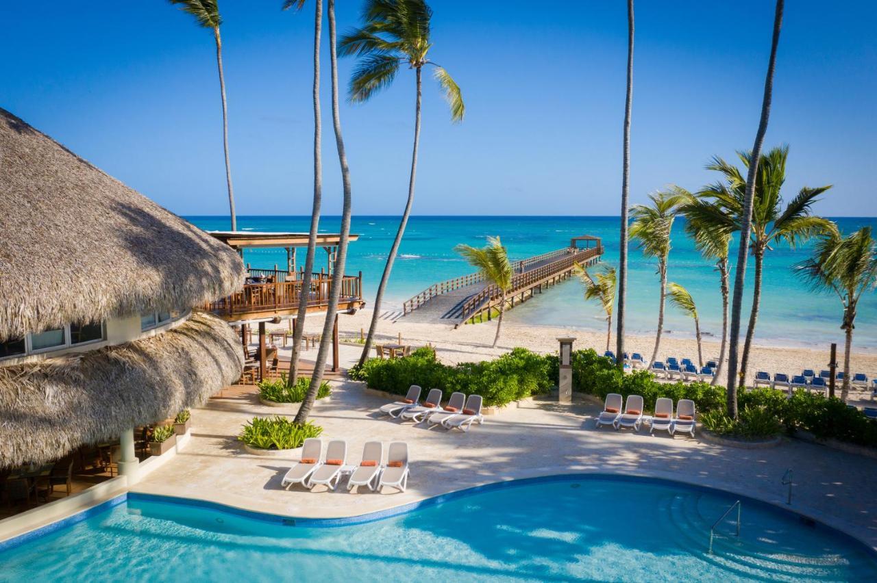 Impressive Punta Cana Esterno foto The photo depicts a serene tropical scene featuring a swimming pool in the foreground, surrounded by lounge chairs. In the background, there is a beautiful beach with clear turquoise waters and a wooden pier extending out into the ocean. Palm trees f
