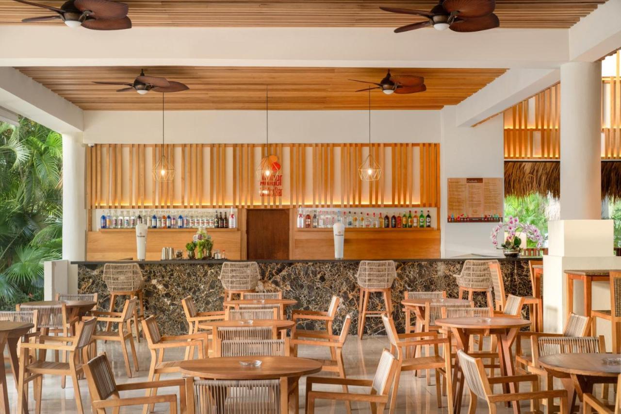 Impressive Punta Cana Esterno foto The photo shows a modern and inviting bar or restaurant interior. It features a spacious layout with wooden flooring and ceiling, complemented by ceiling fans. The bar area is prominently displayed in the background, showcasing a variety of bottles w