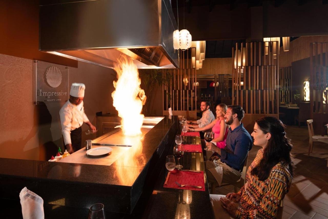 Impressive Punta Cana Esterno foto The photo shows a dining scene in a restaurant setting. A chef in a white uniform is performing a cooking demonstration at a teppanyaki table, creating a large fire as part of the cooking process. In front of the chef, several diners are sitting at t