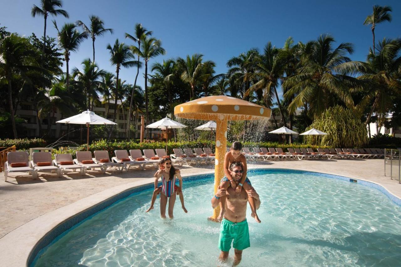 Impressive Punta Cana Esterno foto The photo shows a lively pool scene under a clear blue sky. In the foreground, there are two children playing; one is sitting on the shoulders of an adult. They are enjoying the water and the sunny atmosphere. Nearby, there is a water feature resembl